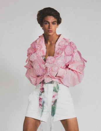 Young adult male poses in a light pink jacket with flower accents and white shorts with a floral belt against a white background.