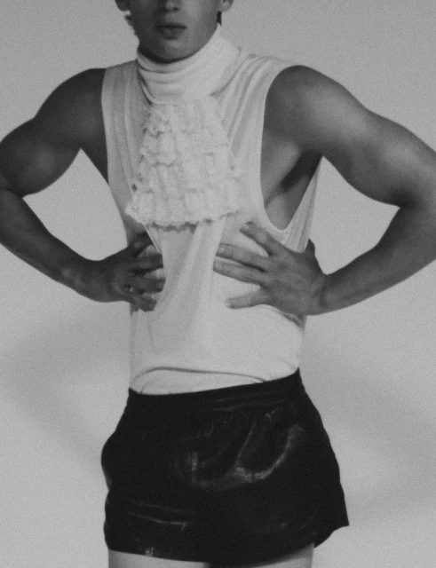 Man in sleeveless top and shorts poses against a white background, showcasing his physique in a monochrome photograph with artistic lighting.