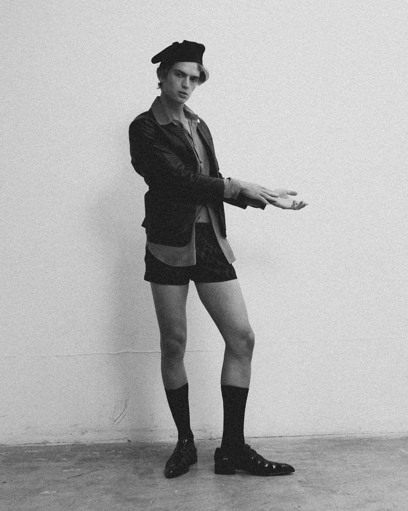 Full-length portrait of a young man in a jacket, shorts, socks, shoes, and beret in a photo studio, posing against a plain wall.