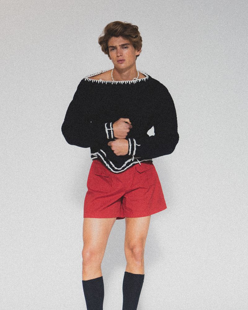 Young adult man poses in a black and white sweater, red shorts, and black socks against a white background in a studio setting.