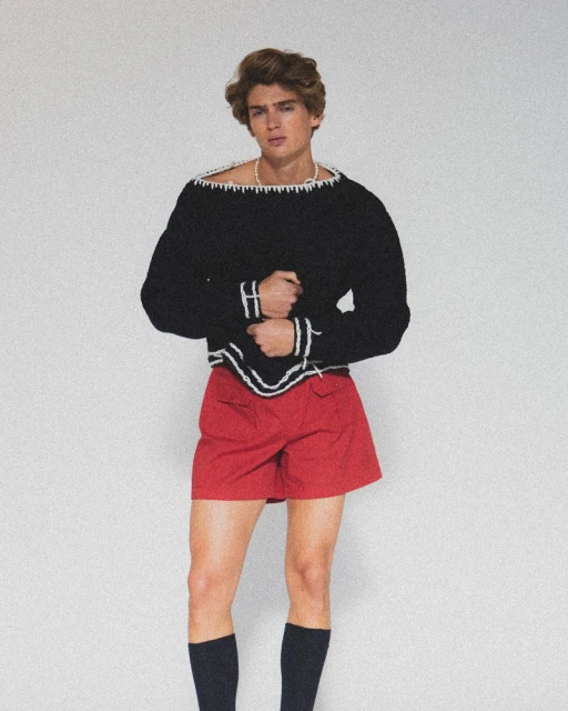 Young adult man poses in a black and white sweater, red shorts, and black socks against a white background in a studio setting.