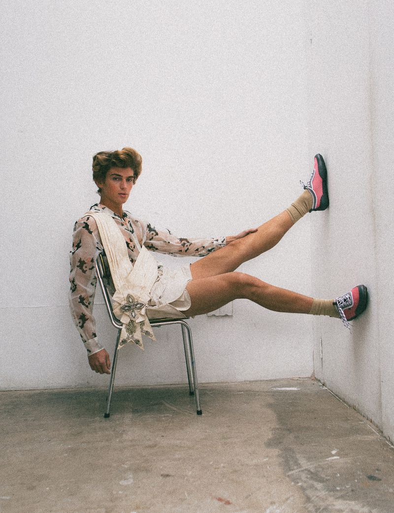 Male fashion model posing in floral shirt and sneakers, seated on metal chair with legs propped against wall, indoor studio shot.