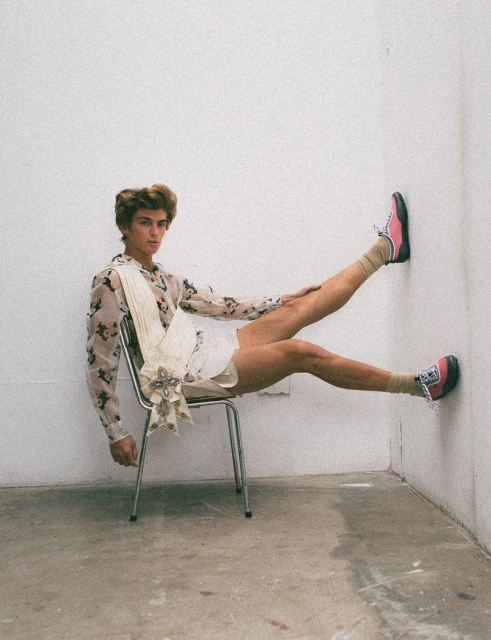 Male fashion model posing in floral shirt and sneakers, seated on metal chair with legs propped against wall, indoor studio shot.