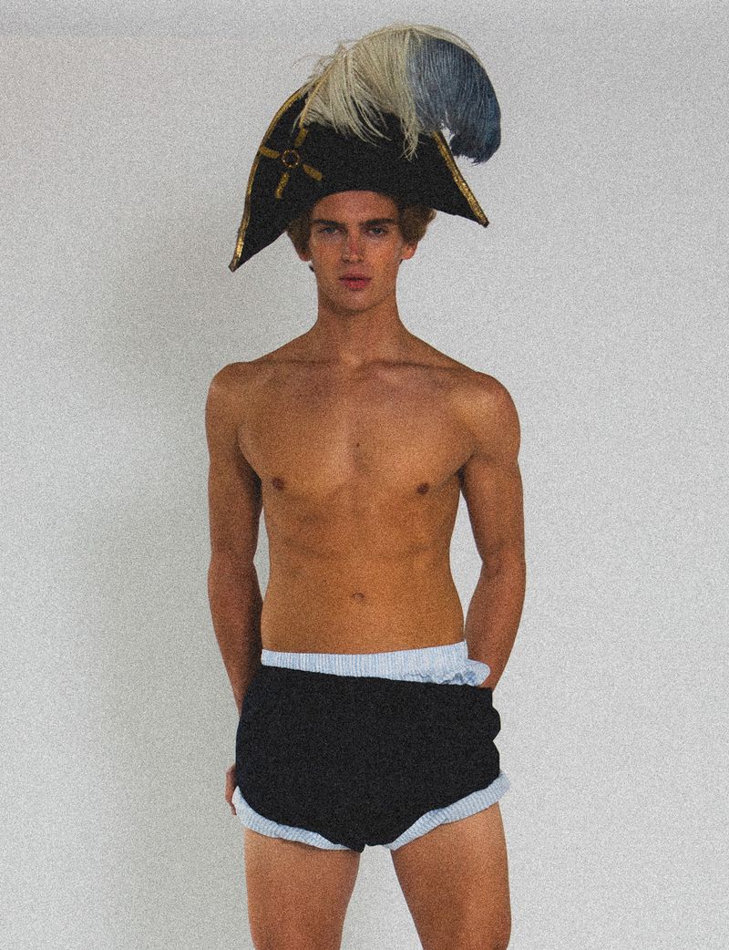 A young, shirtless, light-skinned male model with blond hair poses in black shorts and a costume hat with feathers against a white backdrop.