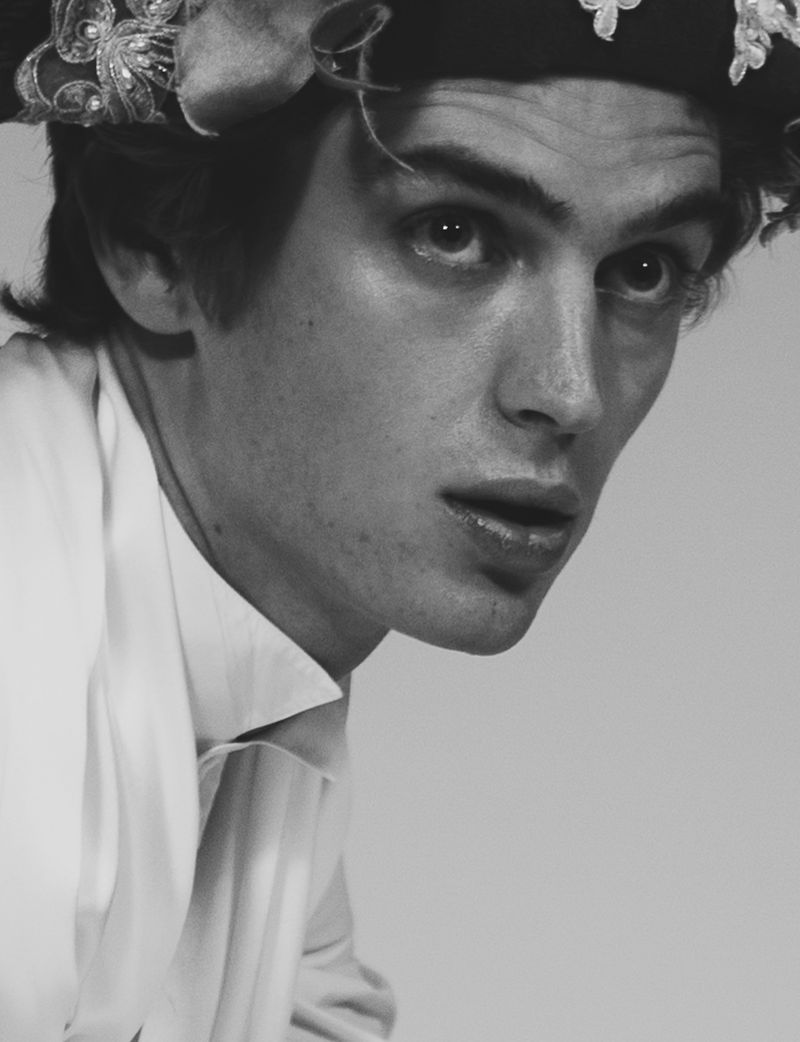 Monochrome portrait of a young adult man with light skin wearing a white shirt and floral headpiece against a light background.