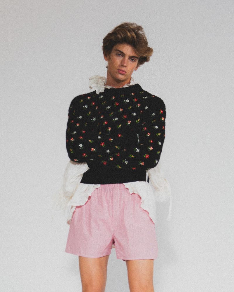 Adult male in a studio, wearing a floral sweater, and pink shorts. He's posing in a full shot, with a white background.