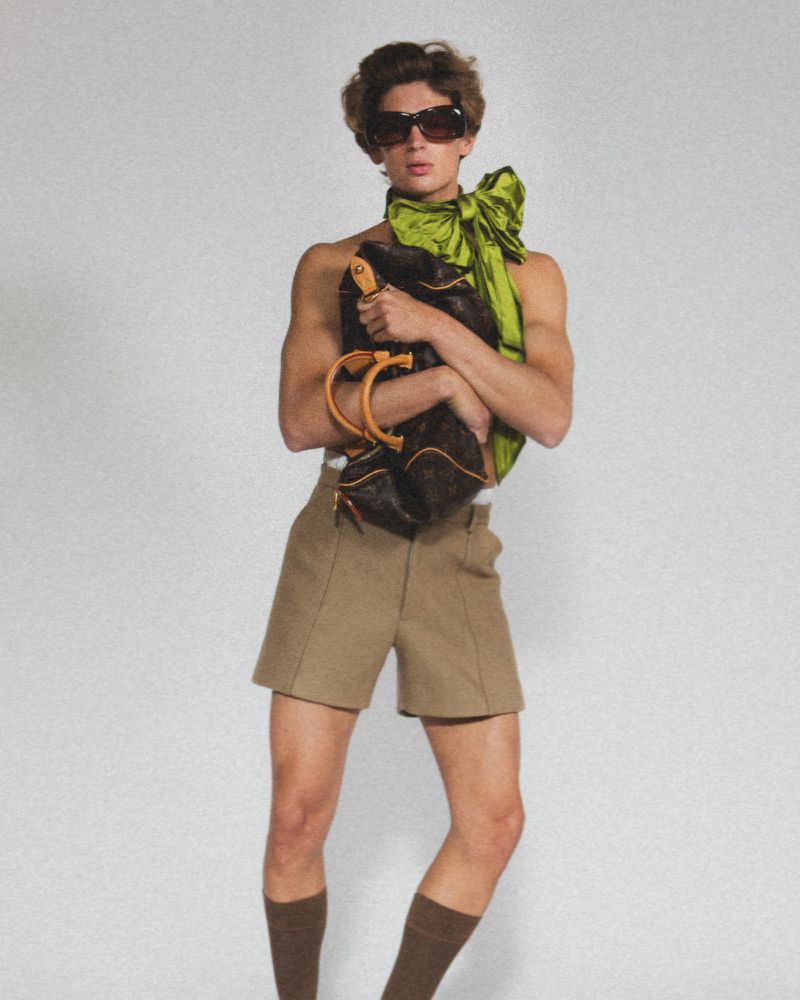 An adult male stands in a photo studio holding a Louis Vuitton handbag, wearing sunglasses, khaki shorts, knee socks, and a green scarf.