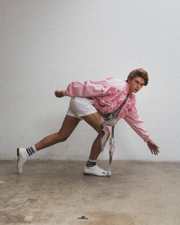 Male model in a pink textured jacket and white shorts posing in a low stance against a plain wall.