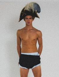 Shirtless male model wearing a black tricorn hat with feathers and dark shorts against a plain background
