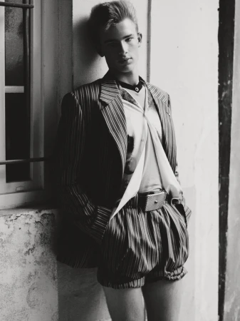Monochrome portrait of a young man in a striped jacket and shorts standing near a window, showcasing retro fashion and style.