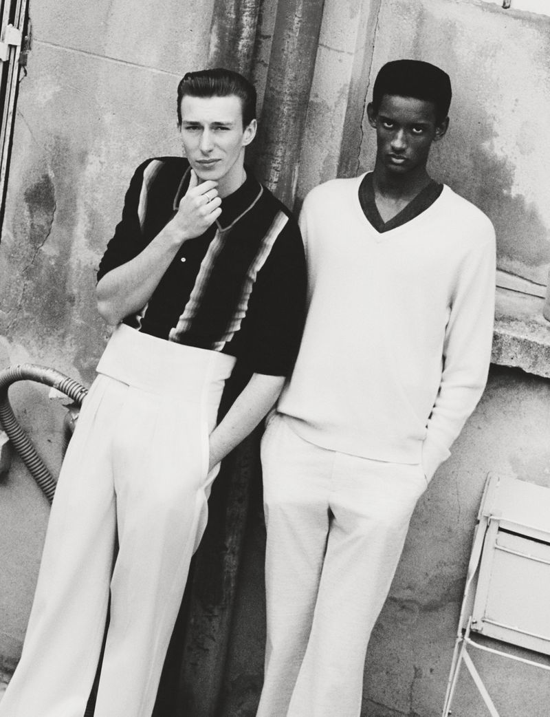 Monochrome portrait of two men in vintage-style attire leaning against a textured wall outdoors, creating a classic fashion editorial image.