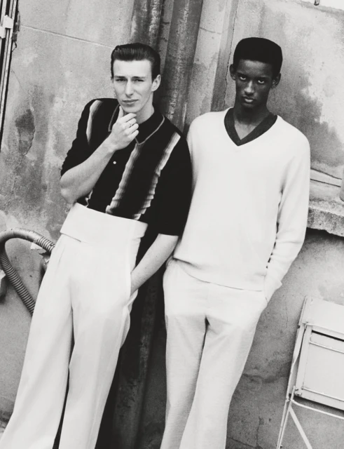 Monochrome portrait of two men in vintage-style attire leaning against a textured wall outdoors, creating a classic fashion editorial image.
