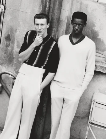 Monochrome portrait of two men in vintage-style attire leaning against a textured wall outdoors, creating a classic fashion editorial image.