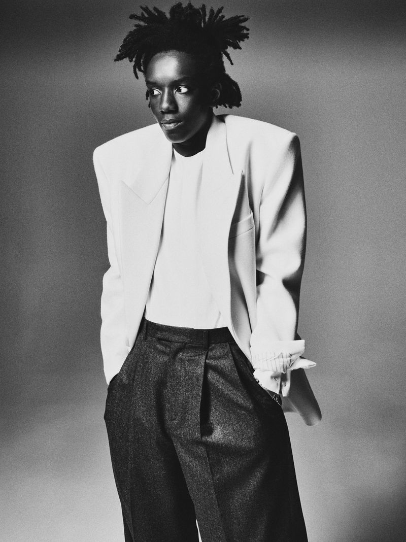 Monochrome portrait of an adult with dreadlocks wearing a white blazer and dark pants against a neutral backdrop.