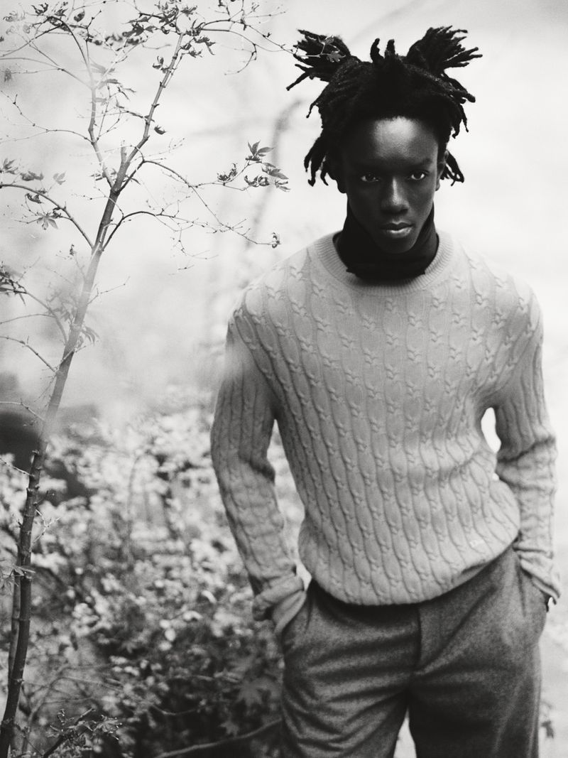Black man wearing a cable-knit sweater standing outdoors near bare trees in a black and white editorial photograph.