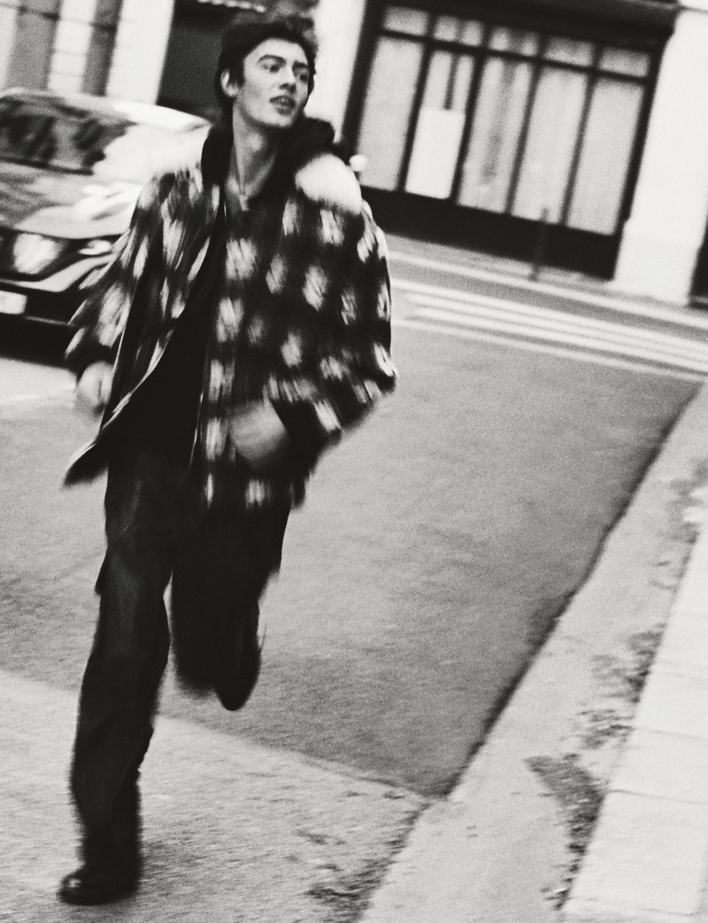 A young adult male in a patterned jacket runs down a city street with buildings and a car visible in the background in black and white.