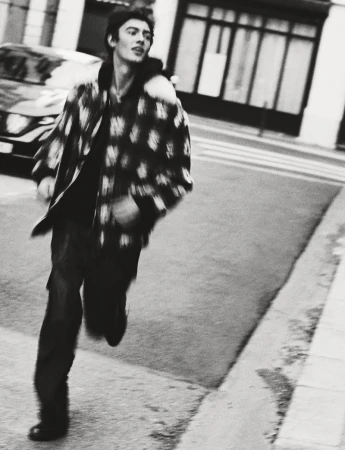 A young adult male in a patterned jacket runs down a city street with buildings and a car visible in the background in black and white.
