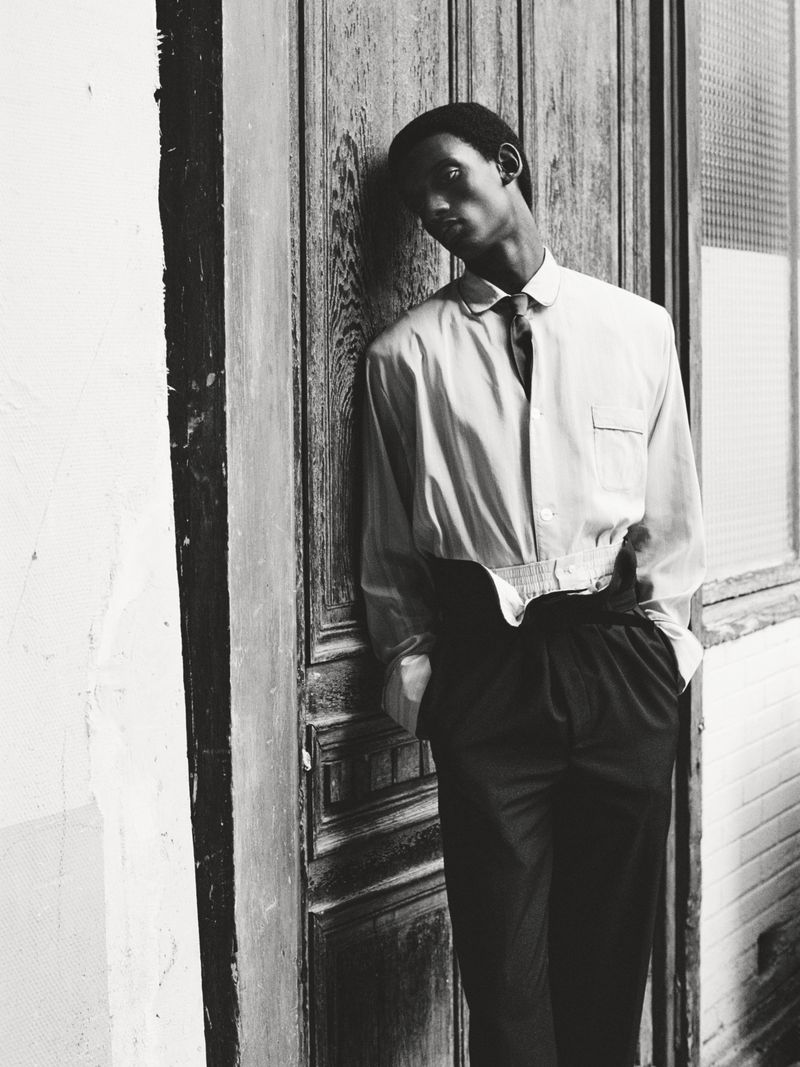 Black and white portrait of a slim African male in dress shirt and tie, leaning against paneled wall with hands in pockets.