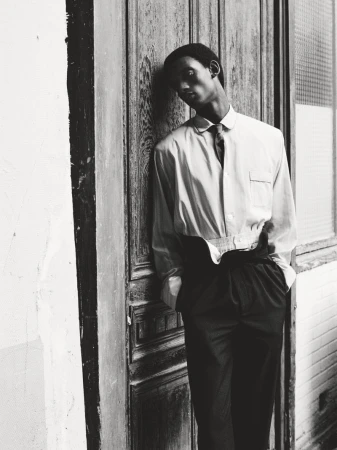 Black and white portrait of a slim African male in dress shirt and tie, leaning against paneled wall with hands in pockets.