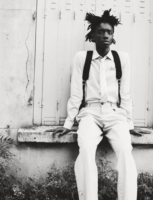 Monochrome portrait of a Black male sitting in front of a boarded-up window wearing a shirt, tie, suspenders, pants, and backpack.