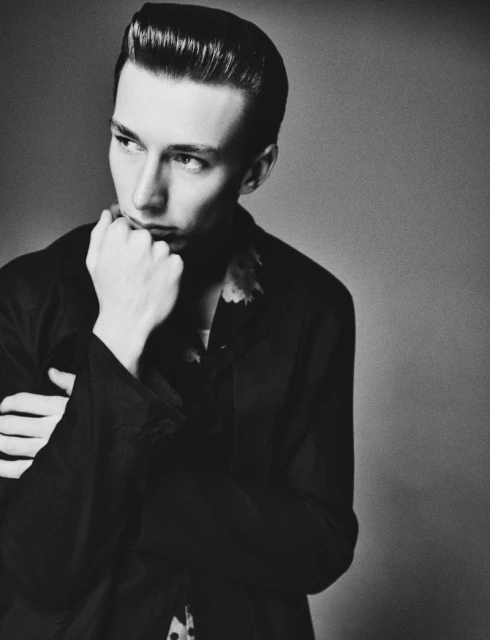 Monochrome portrait of a slender adult male with a pompadour hairstyle, wearing a dark shirt and posing with his hand to his chin.