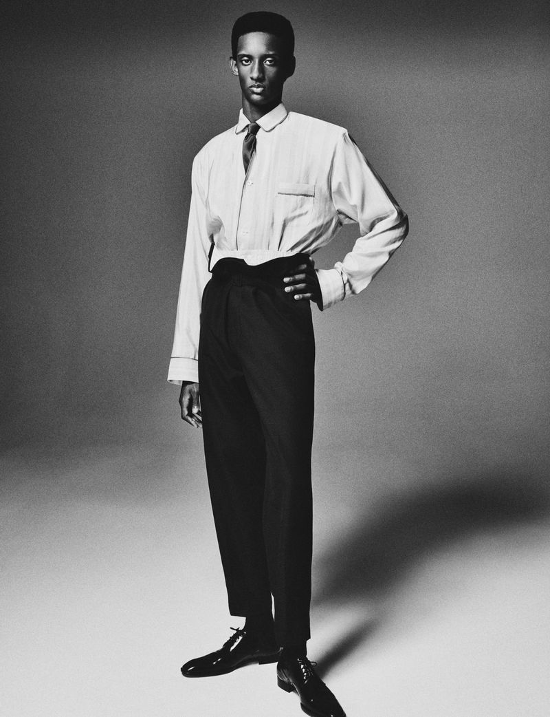 Elegant black male model in a light shirt and black trousers strikes a pose in a full shot, set against a solid gray backdrop.