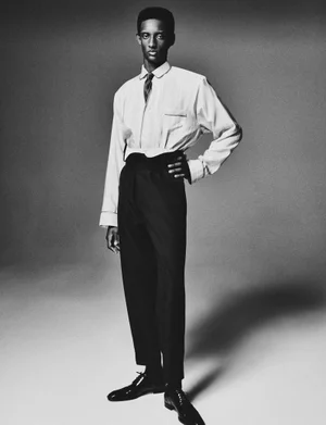 Monochrome portrait of a black male model posing in a white button-down shirt and dark trousers against a grey studio backdrop.