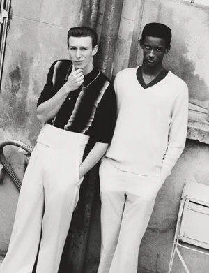 Two male models posing outdoors against a textured stone wall for HERO Magazine, captured in monochrome.