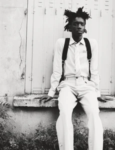 Black and white portrait of a young man seated outdoors wearing a collared shirt, tie, and backpack against a textured wall.