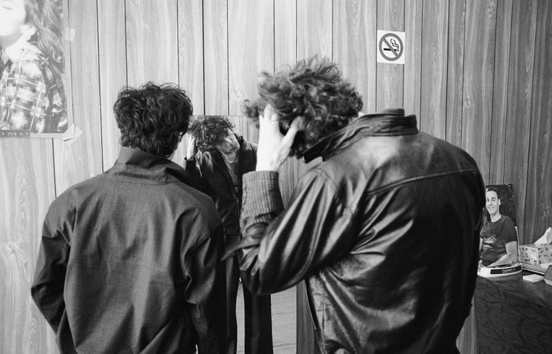 Monochrome shot of three men inside, two looking at the mirror while one man on the far right stands by a table with a photo on it.