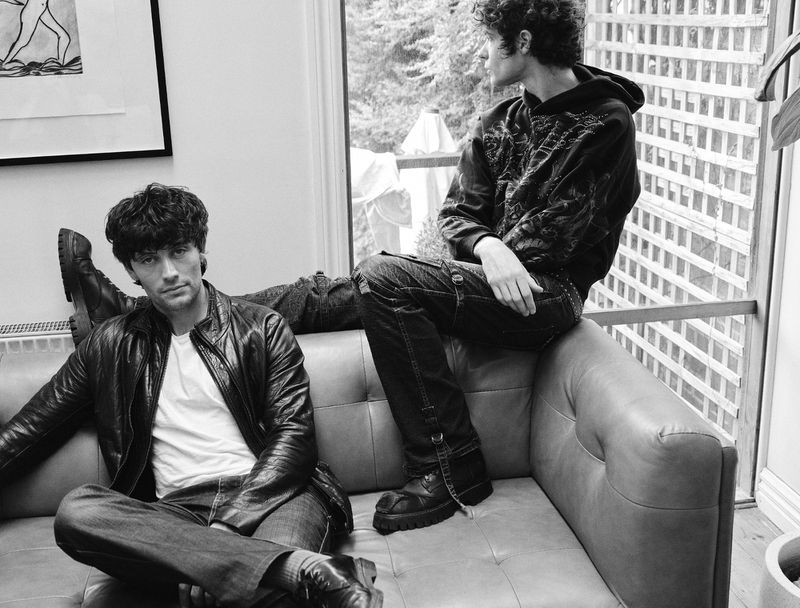 Two adult males pose on a leather couch indoors with natural light. One man sits; the other reclines with legs draped over the couch back.