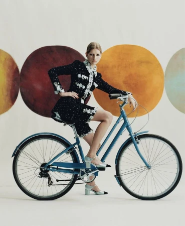Adult female model posing in a black and white dress on a blue bicycle in front of a studio backdrop with colorful circles.