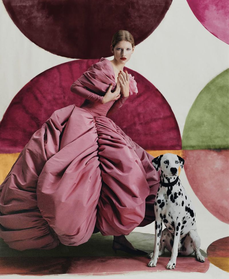 Fashion shot of a woman in a pink dress with a dalmatian dog against an abstract semicircle patterned backdrop in a studio setting.