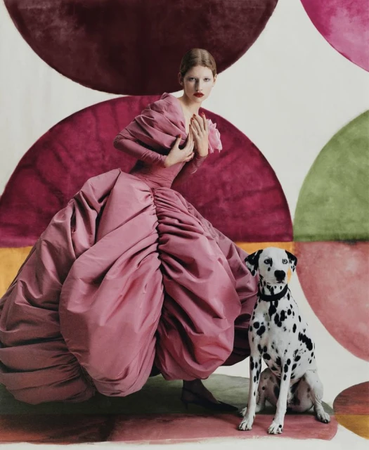 Fashion shot of a woman in a pink dress with a dalmatian dog against an abstract semicircle patterned backdrop in a studio setting.
