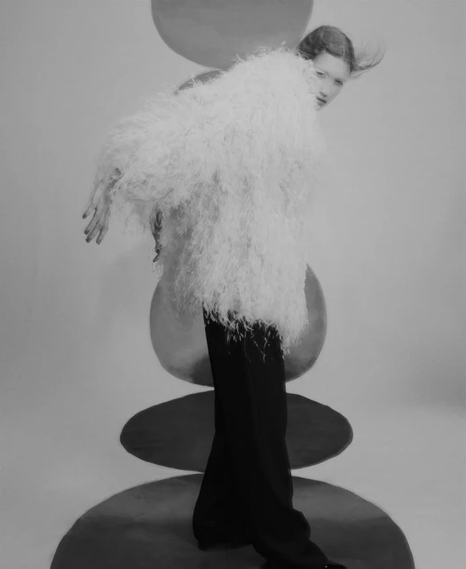 Editorial fashion shot of a woman in a feathered jacket and black pants, posing in front of stacked circular props in black and white.