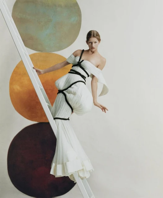 Fashion shot of a blonde adult woman in an off-shoulder white dress with black rope detail climbing a white ladder against a studio backdrop.