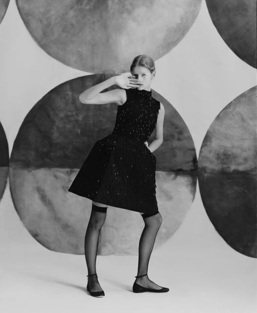 Young woman poses in a black dress with embellished bodice, black stockings, and ballet flats against an abstract circle backdrop.