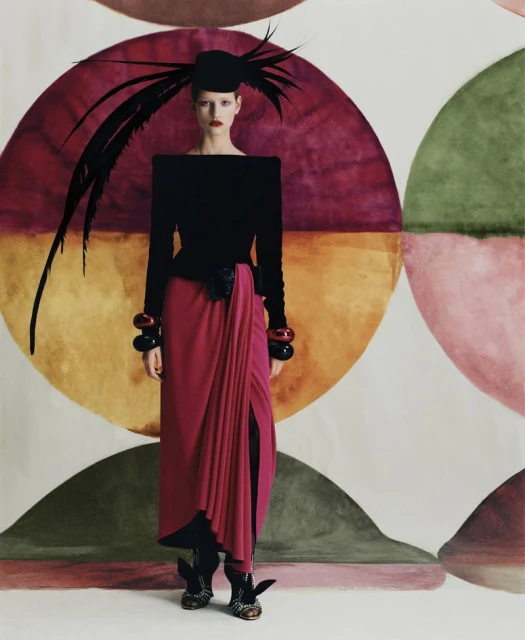 A woman in a black top and pink skirt poses in front of a patterned backdrop, wearing a feathered hat and chunky bracelets, fashion shoot.