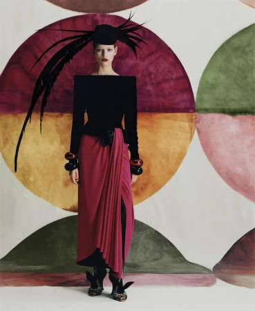 A woman in a black top and pink skirt poses in front of a patterned backdrop, wearing a feathered hat and chunky bracelets, fashion shoot.