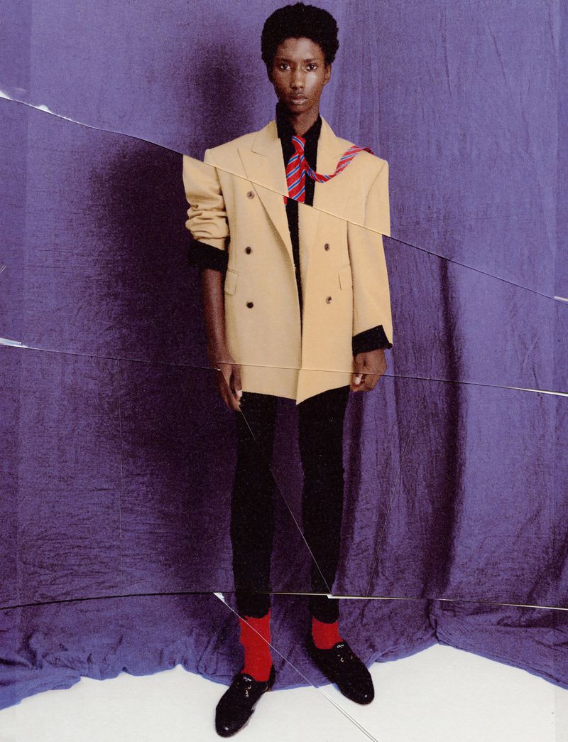 Standing African American model in an oversized tan blazer with a red striped tie, black pants, and red socks poses in a studio.