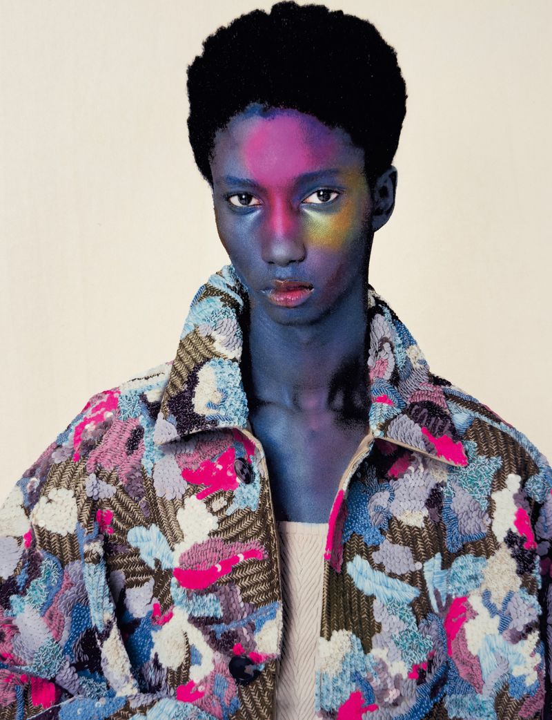 A Black male fashion model with blue skin and colorful face paint wears a patterned jacket in a studio shot, showcasing beauty and style.