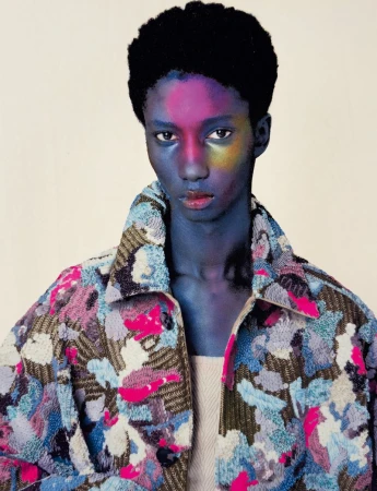 A Black male fashion model with blue skin and colorful face paint wears a patterned jacket in a studio shot, showcasing beauty and style.