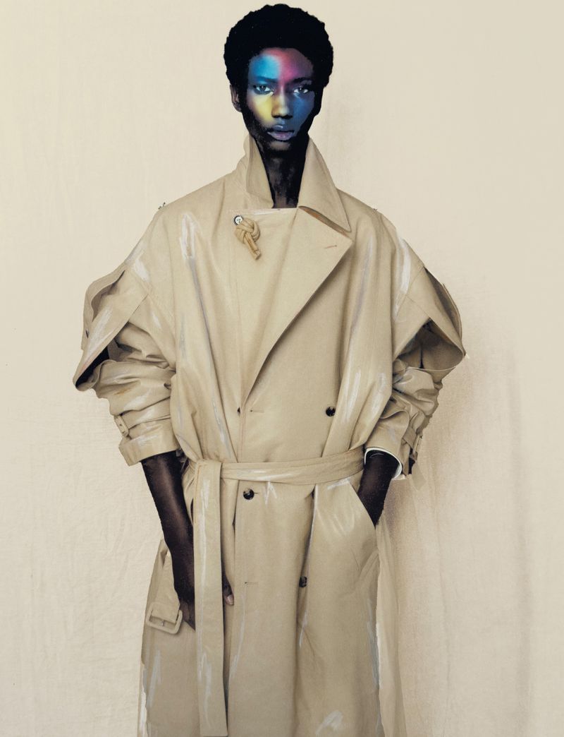 Stylish male model wearing beige trench coat with colorful face makeup in a fashion editorial style studio portrait on neutral backdrop.
