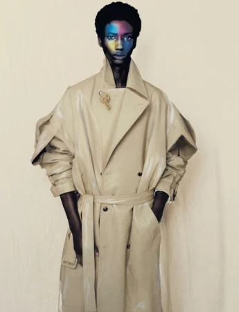 Stylish male model wearing beige trench coat with colorful face makeup in a fashion editorial style studio portrait on neutral backdrop.
