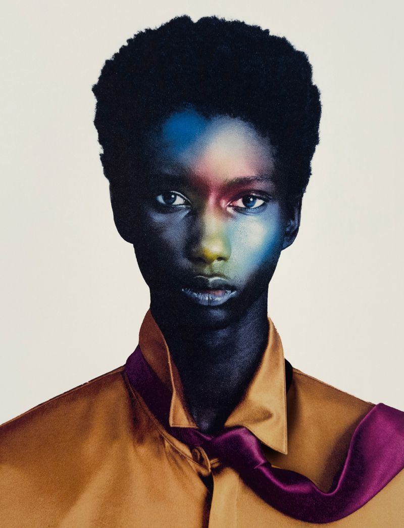 Close-up studio portrait of a young adult Black woman with dark hair, wearing a tan collared shirt and purple necktie against an off-white backdrop.