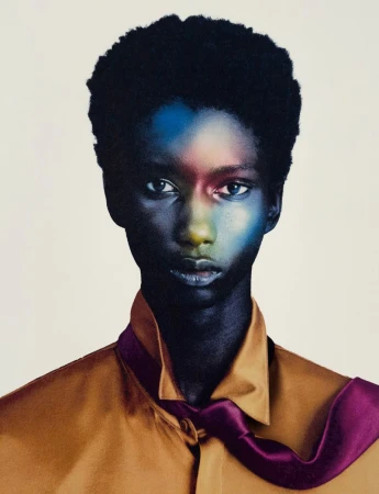 Close-up studio portrait of a young adult Black woman with dark hair, wearing a tan collared shirt and purple necktie against an off-white backdrop.