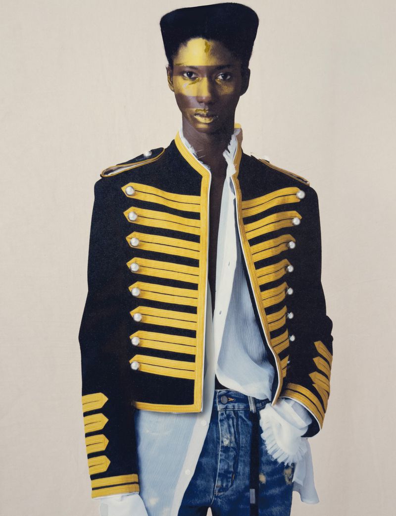A person with gold body paint wears a black and gold military-style jacket, white shirt, blue jeans, and black hat against a cream background.
