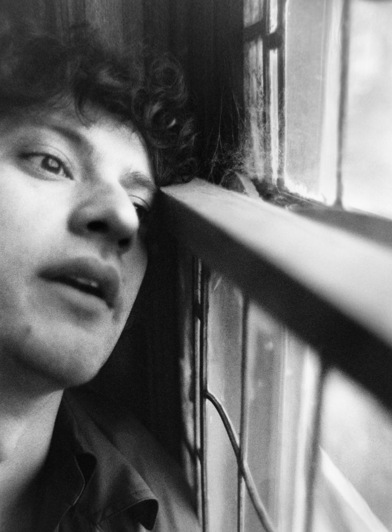 Monochrome portrait of a young adult with curly hair, resting their head against a window frame with a melancholic expression.
