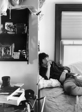 Monochrome portrait of Dominic Sessa reclining on a bed near a window inside a room with shelves and books.