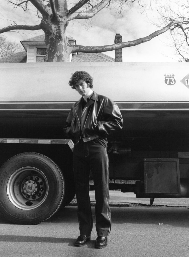 Monochrome portrait of a young adult man in a jacket standing in front of a tanker truck with a tree and house in the background.